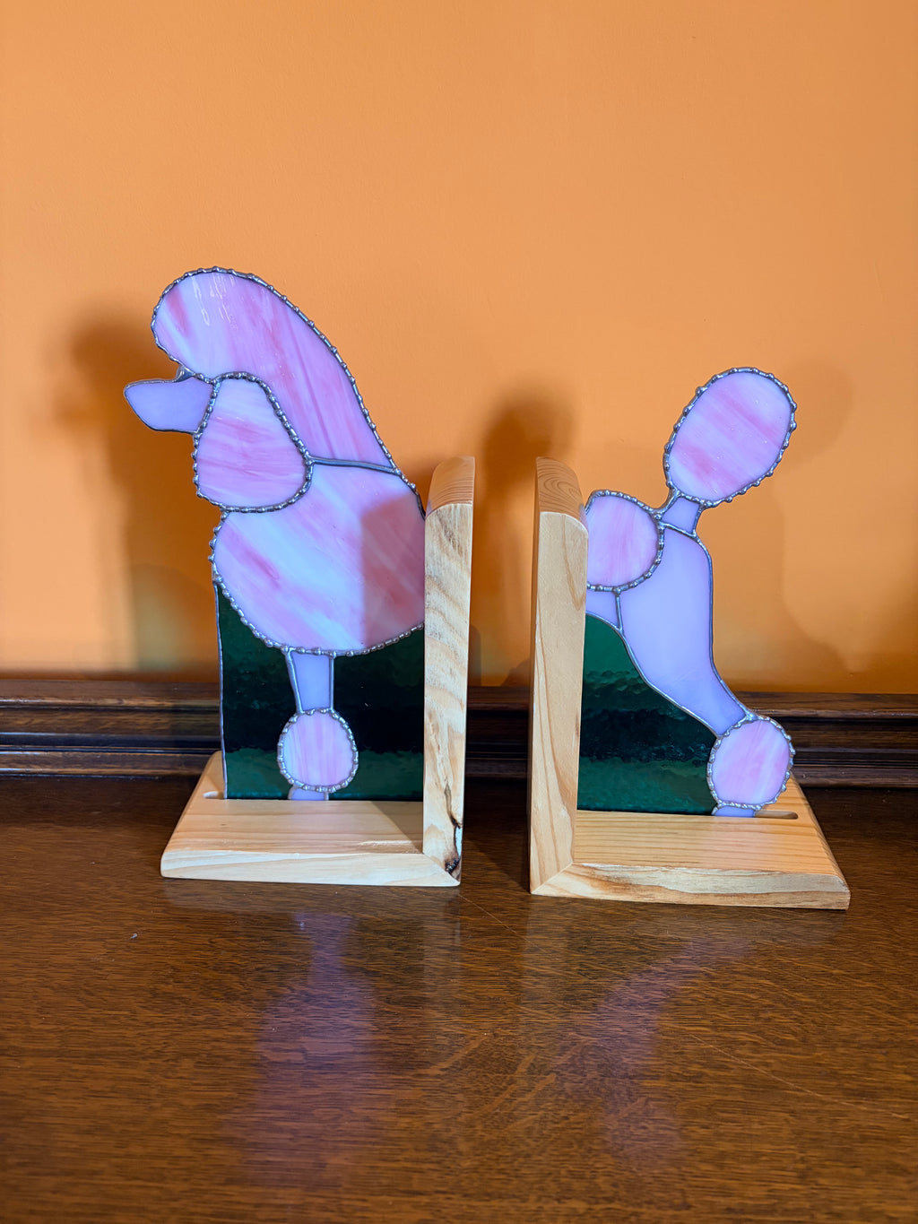 A pair of wooden bookends shaped like poodles, featuring light pink and lavender glass with aqua backing, displayed on a dark wooden table against an orange wall.