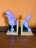 A pair of wooden bookends shaped like poodles, featuring light pink and lavender glass with aqua backing, displayed on a dark wooden table against an orange wall.