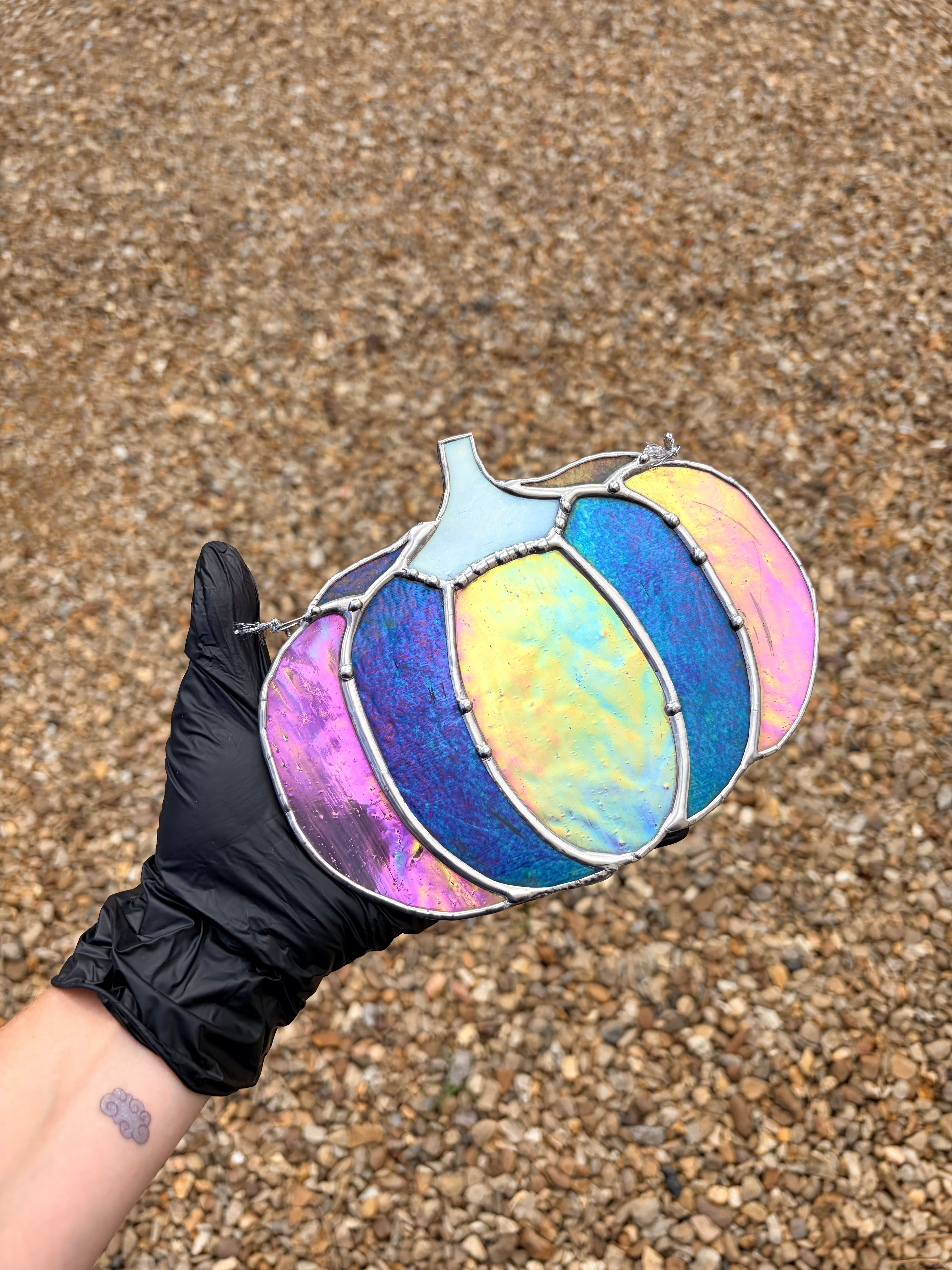 A hand wearing a black glove holds a stained-glass pumpkin ornament with iridescent, multicolored panels in shades of blue, purple, yellow, and pink, set against a background of brown gravel.