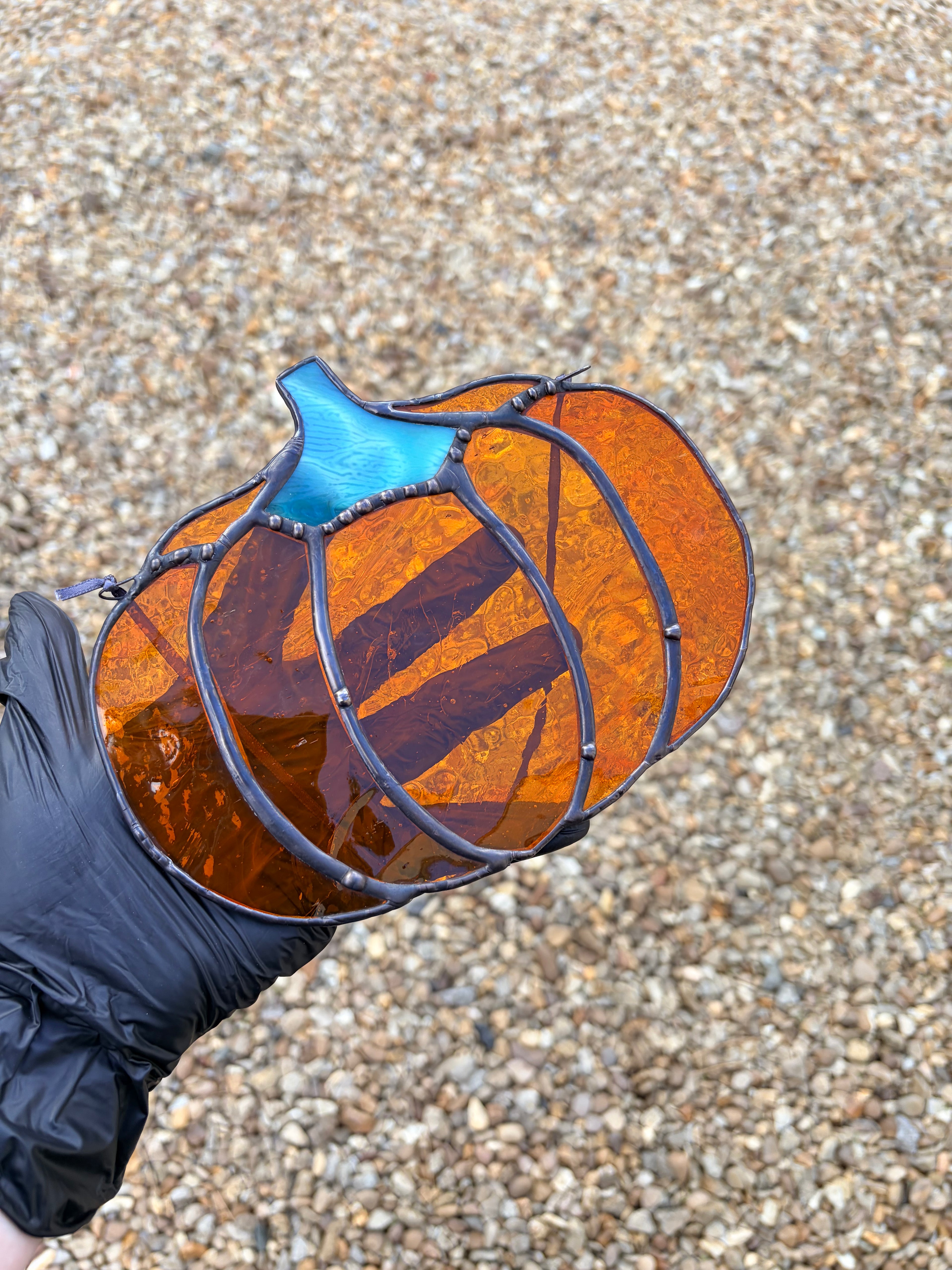 A hand wearing a black glove holds a stained glass pumpkin ornament with orange and blue glass panels and dark metal framing, set against a gravel background.
