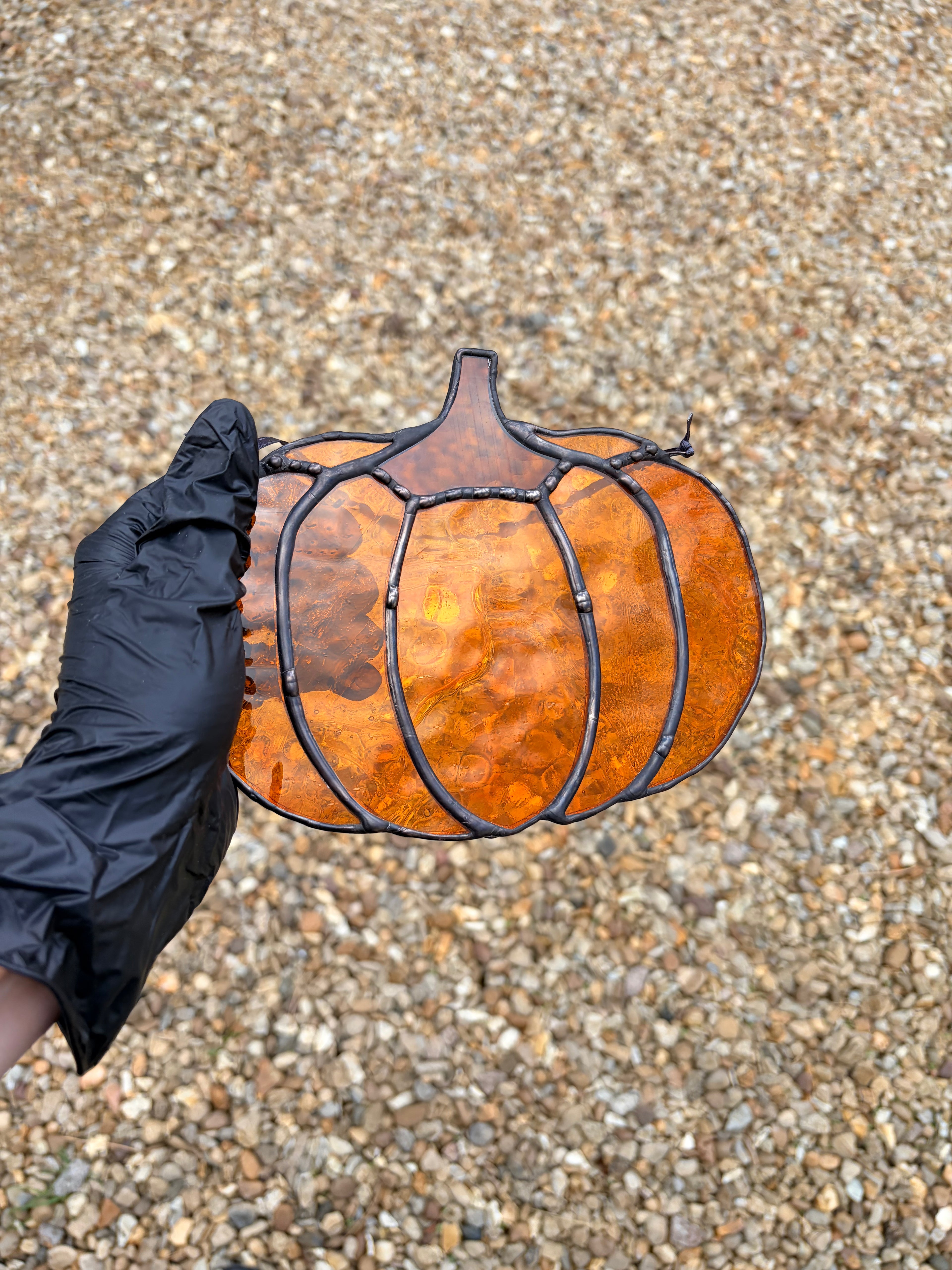 A hand wearing a black glove holds a stained glass pumpkin against a gravel background. The pumpkin features textured orange and amber glass panels divided by black metal frames.