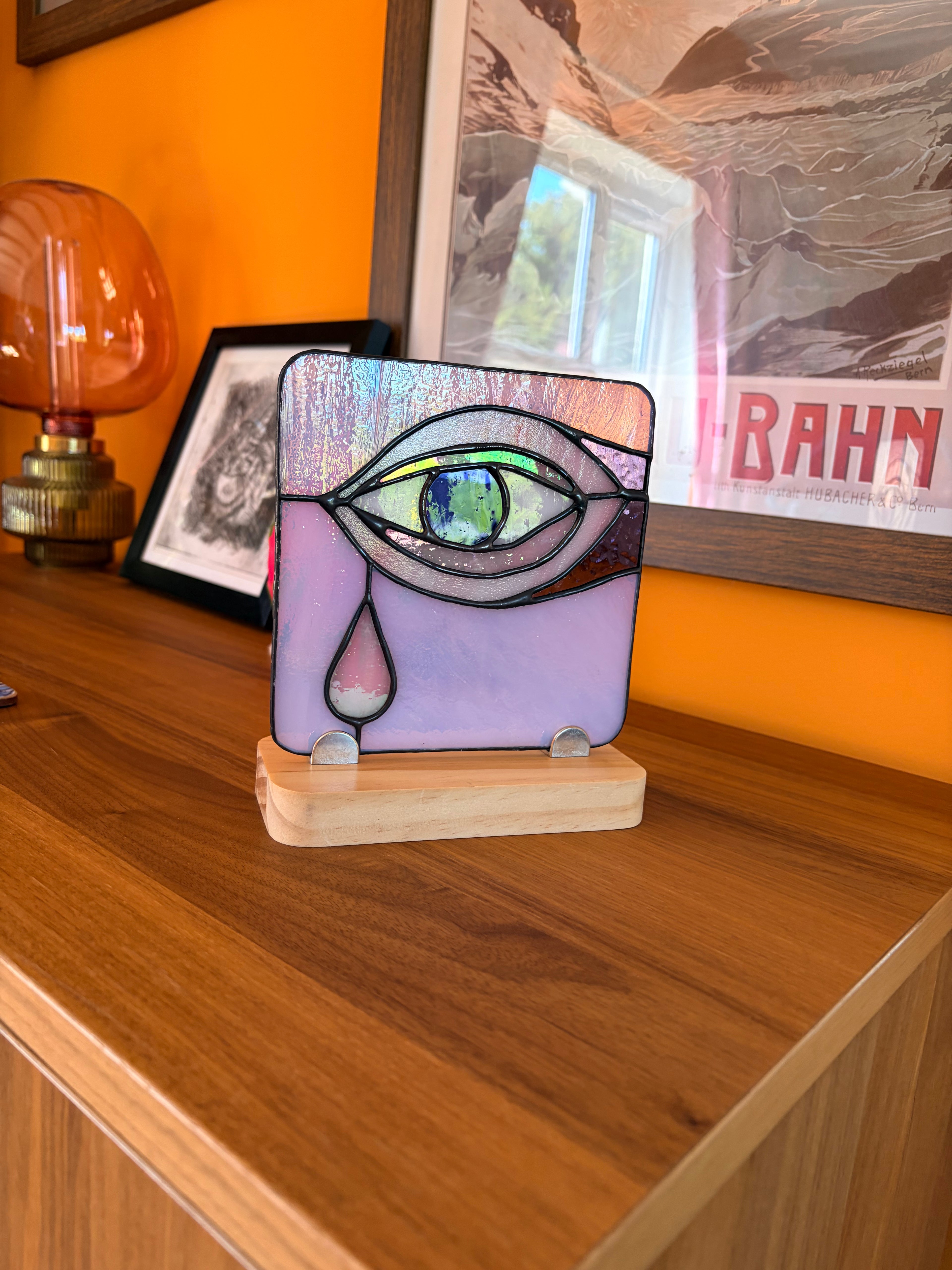 Decorative glass piece with eye design on a wooden surface, with a lamp and framed picture in the background.