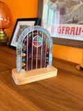 Stained glass fairy door displayed in a wooden stand, lit to show iridescent glass used in the window