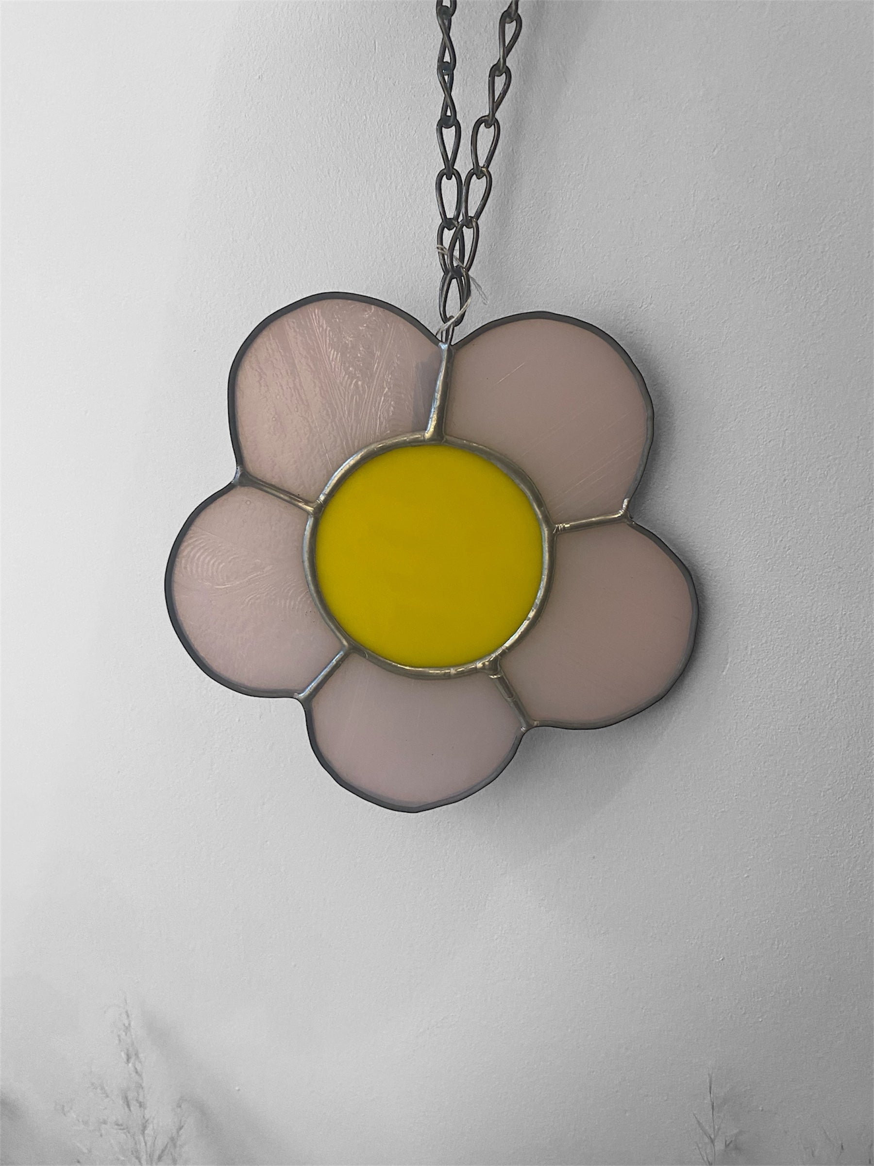 Flower-shaped stained glass suncatcher with yellow centre and pink petals on a chain against a light gray background