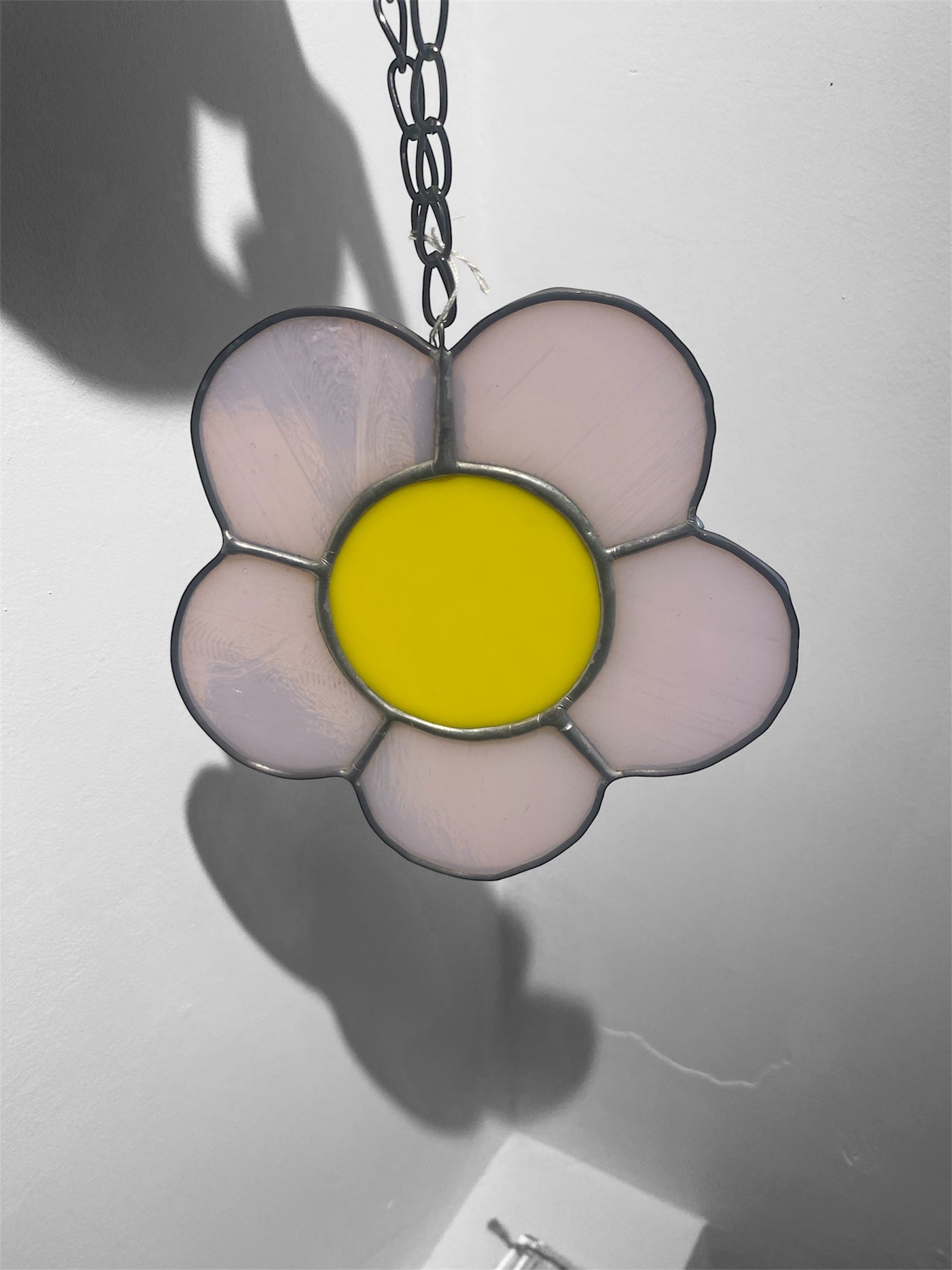 Flower-shaped stained glass suncatcher with yellow centre and pink petals on a chain against a light gray background