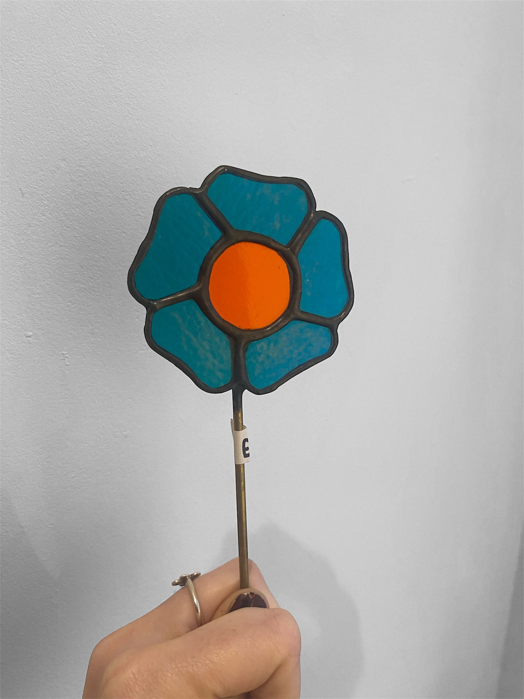 Hand holding a stained glass flower with blue petals and an orange center against a plain background