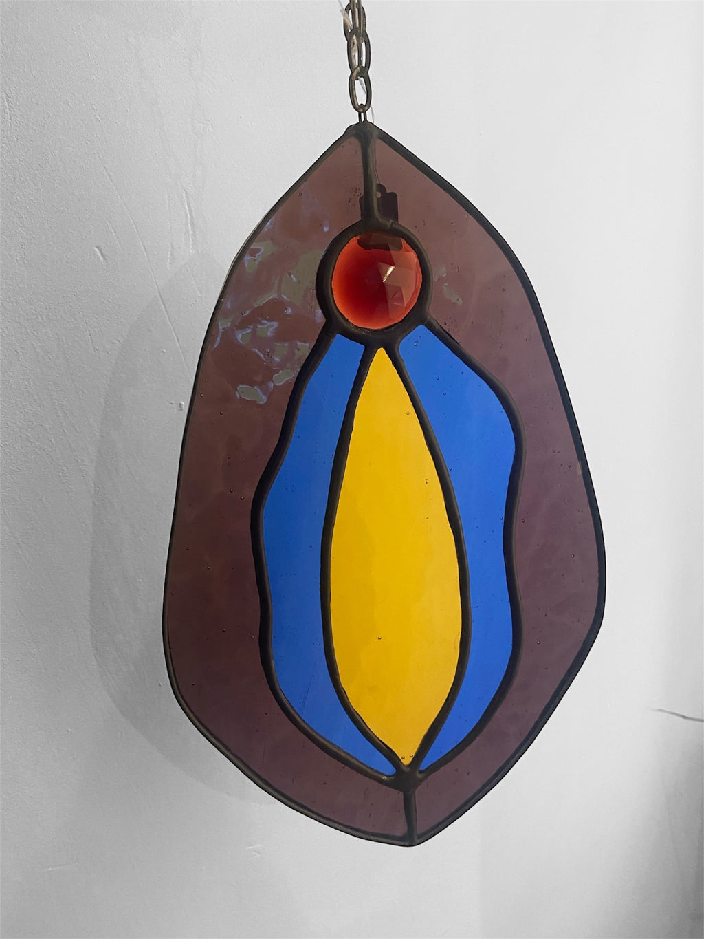 Stained glass hanging panel with blue, purple, pink and yellow vulva design on a plain background
