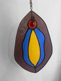 Stained glass hanging panel with blue, purple, pink and yellow vulva design on a plain background