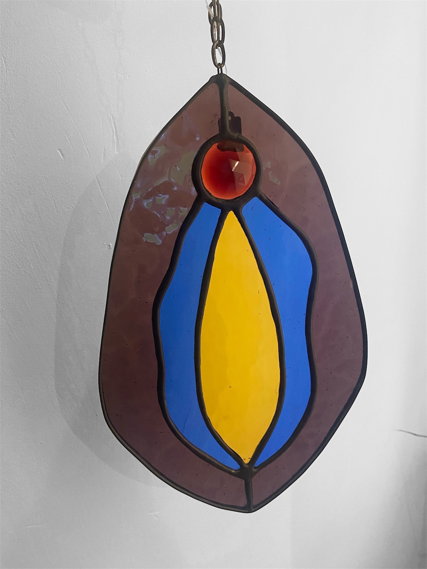 Stained glass hanging panel with blue, purple, pink and yellow vulva design on a plain background