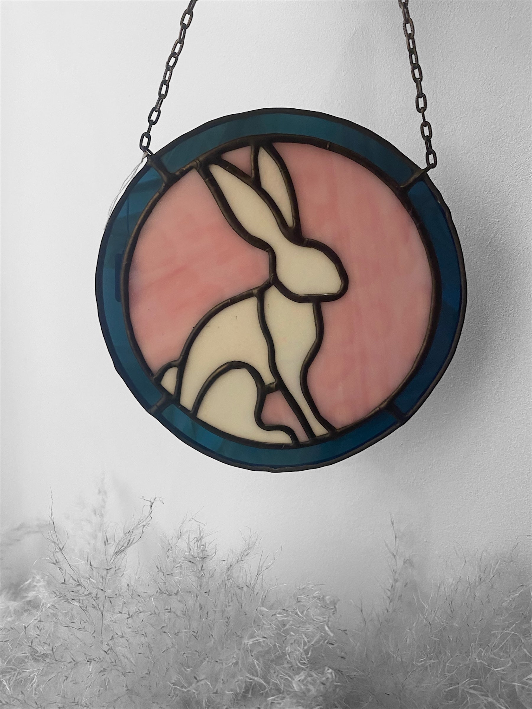 Stained glass window with a rabbit design on a pink background, hanging against a white wall.