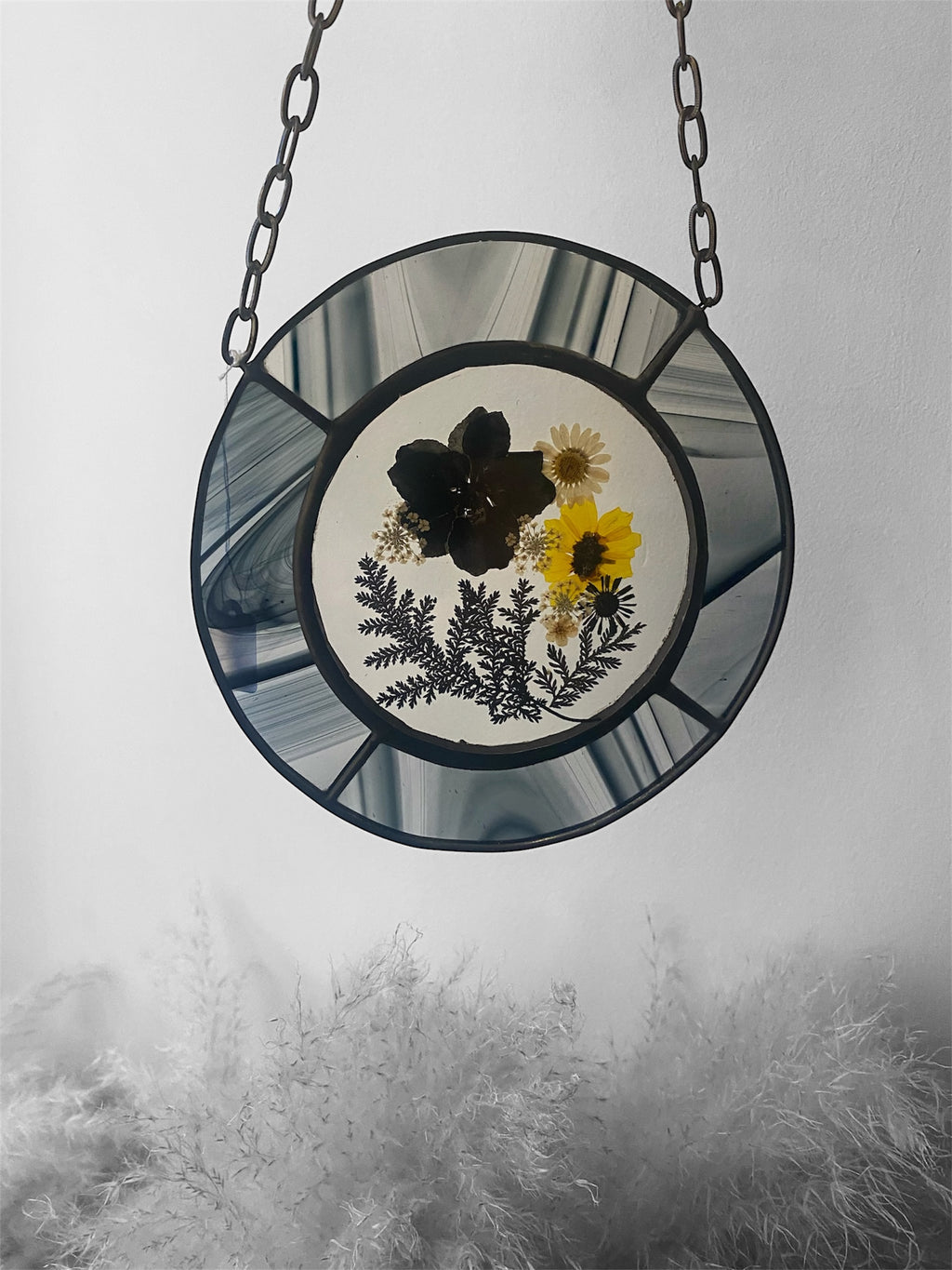 Round stained glass panel with pressed flowers and leaves, hanging against a foggy background.